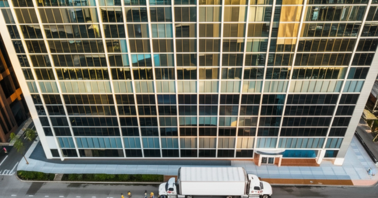 Vista aerea iperrealistica di un edificio per uffici moderno in una città all'alba, con un camion di trasloco e traslocatori caucasici che caricano merci.